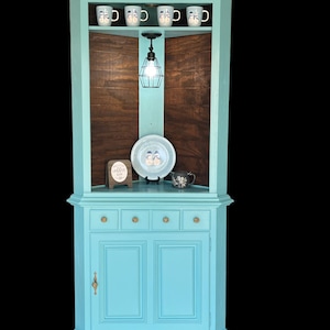 May include: A teal corner cabinet with a wood-paneled back and a light fixture. The cabinet has three drawers and two doors with brass hardware. The top shelf has five mugs and a plate. The cabinet has a sign that says "Coffee is my love language".