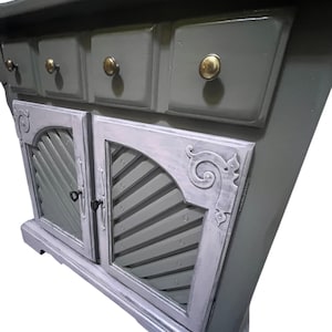 May include: A gray painted wooden cabinet with three drawers and two doors. The drawers have brass knobs. The doors feature a sunburst design and decorative scrollwork. The cabinet has a distressed, vintage look.