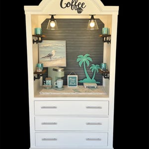 May include: White cabinet with a coffee bar theme. The cabinet has three drawers and a top shelf with a coffee maker, mugs, and a sign that says "Heaven is a cup of coffee and the beach". The top shelf also has a painting of a seagull and two palm trees.
