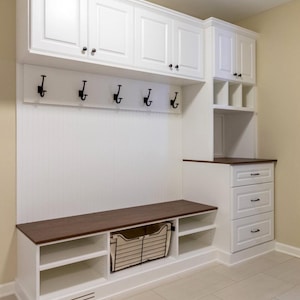 May include: A white entryway storage unit with a dark brown bench top, cabinets, drawers, and open shelving. Black hooks are mounted on the white paneling. A wire basket is placed in the open shelving. The unit is built-in.