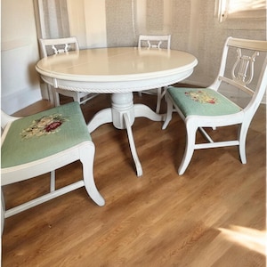 May include: A white round dining table with four matching chairs. The chairs have light green seats with floral embroidery. The table has a pedestal base and a decorative rope trim around the edge. The chairs have a lyre-shaped back.