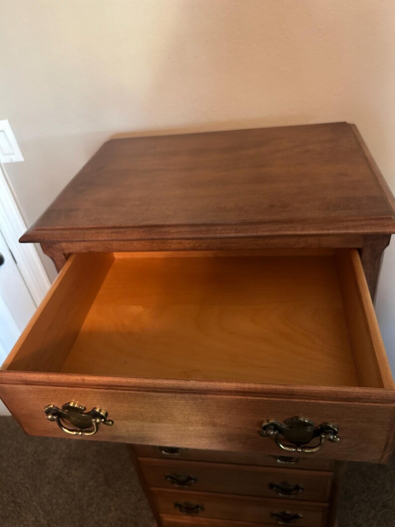 Puede incluir: C&oacute;moda de madera con un caj&oacute;n abierto. La c&oacute;moda es de color marr&oacute;n claro con tiradores dorados. El caj&oacute;n abierto revela el interior. La veta de la madera es visible.