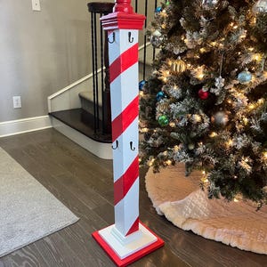 May include: A tall, decorative Christmas coat rack painted in red and white stripes, resembling a candy cane. The rack has a red top and base, with black hooks for hanging items. A Christmas tree with ornaments is in the background.