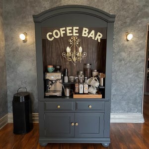 May include: A dark gray coffee bar cabinet with the words "COFFEE BAR" in illuminated letters. The cabinet features a built-in espresso machine, coffee cups, and various coffee-related accessories. A small chandelier hangs inside the cabinet.