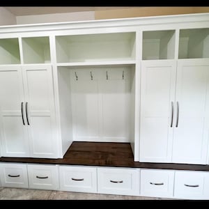 May include: A custom-built white storage unit with multiple compartments. It features upper cabinets, open shelves, and lower drawers. The unit includes a bench with a dark wood top and is designed for entryway or mudroom organization.