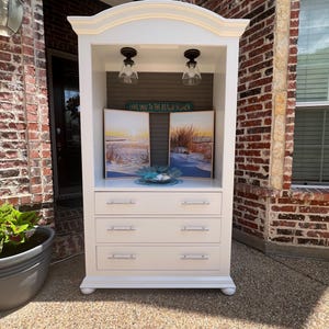 May include: A white cabinet with three drawers and a display shelf. The shelf holds two framed beach landscape paintings, a decorative blue seashell, and a sign that reads "Love You To The Beach & Back". Two light fixtures are mounted above the shelf.