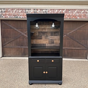 May include: A black cabinet with a wooden interior and two small pendant lights. The cabinet has two drawers with gold knobs and two doors with gold knobs. The cabinet is painted black with a gray base.