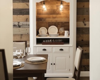 May include: A white wooden hutch featuring two drawers and two lower cabinet doors, with an open upper shelf displaying white plates, bowls, a pitcher, and glassware. Two pendant lights hang inside the hutch, which is set against a wood plank wall.