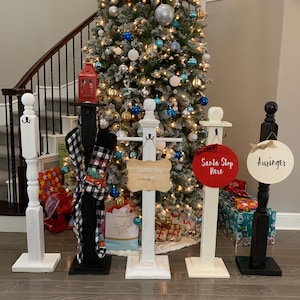 May include: Four wooden Christmas stocking holders with signs. The signs read "Santa Stop Here" and "Auringer". The holders are painted white and black. The holders are in front of a Christmas tree with blue and silver ornaments.