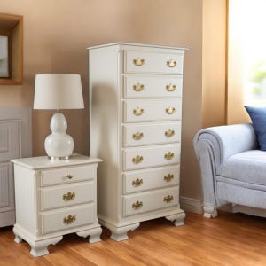Puede incluir: Un juego de muebles de dormitorio blancos con una c&oacute;moda alta y una mesita de noche. Ambas piezas tienen herrajes dorados y tiradores decorativos. Una l&aacute;mpara blanca con una pantalla se encuentra sobre la mesita de noche.