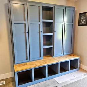 May include: A custom-built, blue storage unit with multiple cabinets and open cubbies. The unit features a natural wood bench seat and a series of closed cabinets above. The design is ideal for entryways or mudrooms.