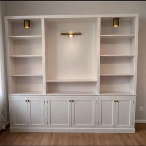 May include: A large, white built-in cabinet with multiple shelves and cabinets. The cabinet has three sections, with shelves on either side and a central section with a shelf and a light fixture. Gold-colored lights are mounted above the shelves.