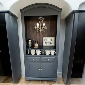 May include: A dark gray coffee station with a wooden interior, featuring a small chandelier, coffee mugs, and a sign that reads "Coffee Lover." The cabinet has drawers and doors, and is flanked by dark gray cabinets.