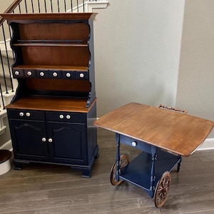 May include: A dark blue hutch with a wood top and shelves, and a dark blue rolling cart with a wood top and a drawer. Both pieces of furniture have white knobs.