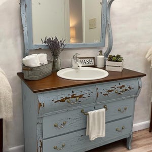 May include: A distressed blue vanity with a wooden countertop and a white oval sink. The vanity has three drawers with brass hardware and a matching mirror. A small sign reads "VASH". The vanity is decorated with a vase of lavender, white towels, and a small plant.