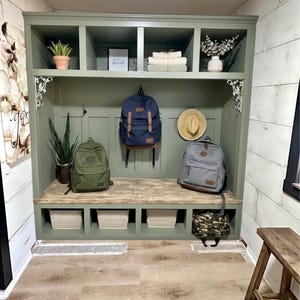 May include: A sage green entryway storage unit with a bench, cubbies, and upper shelves. It features backpacks, a hat, and decorative items. The unit includes three woven baskets and a camouflage bag.