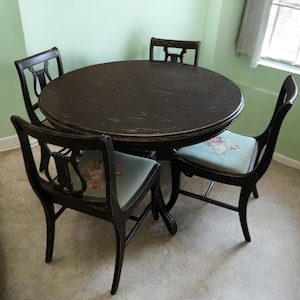 May include: A round, dark brown dining table with four matching chairs. The chairs have black frames and light blue seats with floral patterns. The table and chairs are set against a light green wall.