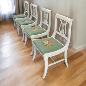 May include: Four white wooden chairs with a lyre-shaped backrest design. Each chair has a green seat cushion adorned with a floral pattern. The chairs are arranged in a row on a hardwood floor, creating a classic and elegant aesthetic.