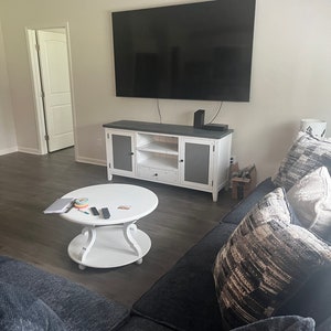 May include: A white entertainment center with a black TV mounted above it. The entertainment center has two doors, two shelves, and a drawer. A black couch with pillows sits in front of the entertainment center. A white coffee table with a laptop, remote controls, and other items sits in front of the couch.