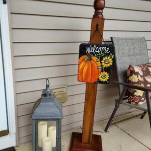 May include: A wooden sign post with a welcome sign featuring a pumpkin and sunflowers. The sign is hanging from a hook on the post. A gray lantern with three candles is on the left side of the image.