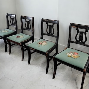 May include: Four antique black chairs with a distressed finish. Each chair features a lyre-shaped back and a square seat upholstered in a light green fabric with a floral design. The chairs are arranged in a row against a white wall.
