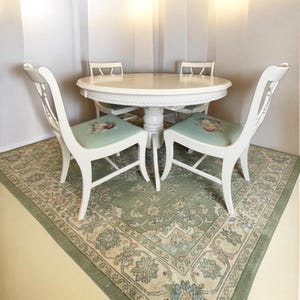 May include: A white round dining table with four matching chairs. The chairs have floral patterned seat cushions and curved backs. The table and chairs are on a green and beige patterned rug.