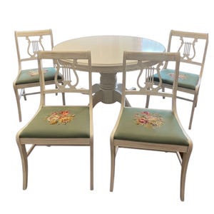 May include: A white round dining table with four matching chairs. The chairs have a lyre-shaped back and green upholstered seats with floral embroidery. The table and chairs are painted in a light, neutral color.