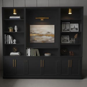 May include: A large, dark charcoal gray built-in wall unit featuring multiple open shelves and lower cabinets with vertical gold handles. The unit includes integrated gold lighting, a central abstract painting, and various decorative items like books, framed photos, and a clock.
