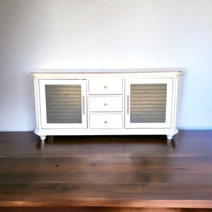 May include: White wooden cabinet with three drawers and two glass doors. The cabinet has a distressed finish and gold-toned hardware.