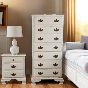 Puede incluir: Un juego de dormitorio blanco con una c&oacute;moda alta y una mesita de noche. Los muebles tienen un acabado envejecido con herrajes de color bronce. Una l&aacute;mpara blanca est&aacute; sobre la mesita de noche, y una cama es parcialmente visible.