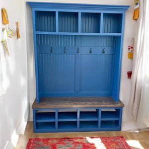 May include: A blue wooden entryway bench with a built-in shelf and hooks. The bench has a dark wood seat and a series of cubbies below. The back of the bench features vertical paneling and open shelves at the top. A red rug is on the floor.