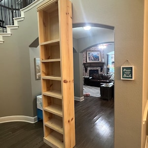 May include: Tall, unfinished wooden bookshelf with multiple shelves. The bookshelf is made of light-colored wood and stands upright against a wall. The shelves are open and ready for storage or display.