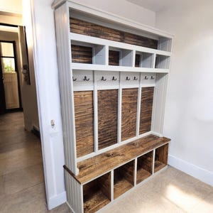 May include: A white and wood entryway organizer with a bench and storage cubbies. The unit features a top shelf with cubbies, a row of hooks, and vertical wood panels. The bench has open storage below. The unit is made of wood.