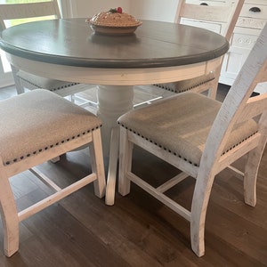 May include: A white farmhouse style dining table with a gray top and four chairs. The chairs have a white frame and beige fabric seats. A pie dish with a red cherry on top sits on the table.