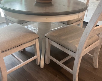 May include: A white farmhouse style dining table with a gray top and four chairs. The chairs have a white frame and beige fabric seats. A pie dish with a red cherry on top sits on the table.