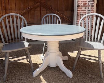 May include: A white pedestal table with a gray top and three gray chairs. The table and chairs are in a rustic setting.