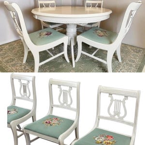 May include: A white dining set featuring a round table and four chairs. The chairs have a lyre-shaped back and floral patterned seat cushions. The table and chairs are painted white, creating a cohesive and elegant look. The table is approximately 48 inches in diameter.