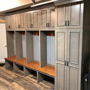 May include: A custom-built storage unit with multiple compartments. The unit features a series of cabinets with black handles, open locker spaces with wooden benches, and a tall cabinet on the right. The wood has a gray stain.