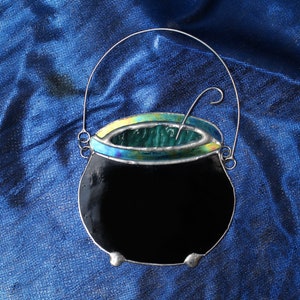 May include: A black stained glass cauldron with a green liquid inside and a silver handle. The cauldron is hanging from a silver wire.