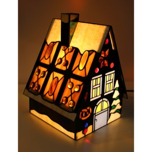 May include: A stained glass lamp shaped like a gingerbread house with a snowy roof, lit from within, with a black chimney and a small tree on the side.