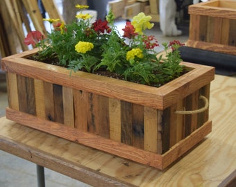 Op de afbeelding: Een rechthoekige houten plantenbak met een touwhandvat. De plantenbak is gemaakt van gerecycled hout met verschillende kleuren en texturen. Hij is gevuld met verschillende kleurrijke bloemen, waaronder gele, rode en oranje.