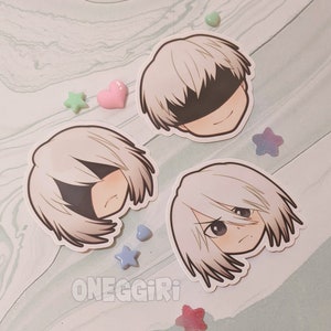 May include: Three white and gray cartoon character stickers with black eyes and hair. The stickers are on a light gray background with pink, green, and blue star-shaped stickers.
