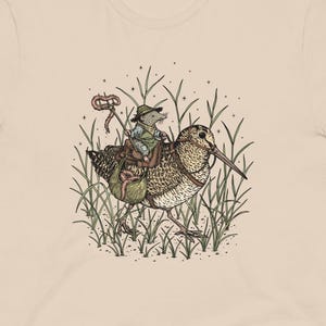 May include: A beige t-shirt with a detailed cartoon illustration. The graphic shows a mouse in a hat and jacket riding a bird through grass. The bird has brown and tan feathers. The mouse is holding a rope. The design is in a detailed, cartoon style.