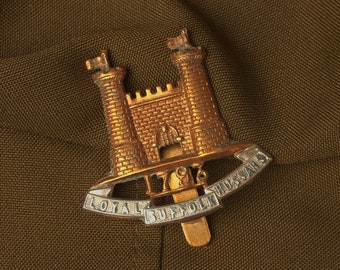 May include: A gold-colored metal cap badge with a castle design and the text "LOYAL SUFFOLK HUSSARS 1793" on a brown fabric background.
