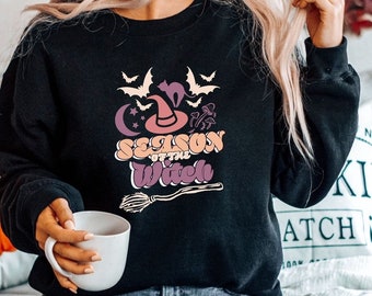 Op de afbeelding: Zwart sweatshirt met een grafische print van een heksenhoed, vleermuizen, paddenstoelen en de tekst "Season of the Witch".