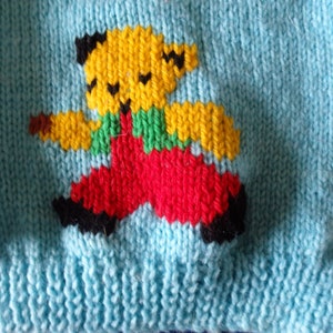 May include: A light blue knitted sweater featuring a yellow teddy bear design with black accents, green overalls, and red pants. The sweater is made of a soft, textured yarn, perfect for children's apparel.