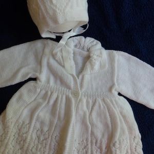 May include: White knitted baby cardigan and matching bonnet with a delicate lace pattern. The cardigan has a button closure and a ruffled collar.