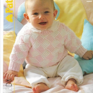 May include: A baby wearing a pink and white checkered sweater with a white collar and white pants. The sweater is part of a knitting pattern. The baby is sitting on a bed with colorful pillows.