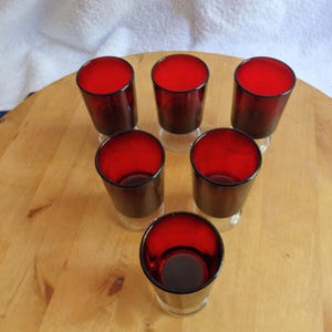 Puede incluir: Conjunto de seis vasos de chupito de vidrio rojo con base transparente. Los vasos están dispuestos en círculo sobre una superficie de madera.
