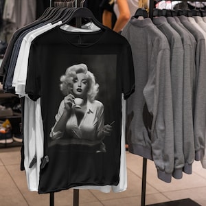 May include: Black t-shirt featuring a black and white image of Marilyn Monroe holding a cup and a cigarette. The t-shirt is displayed on a clothing rack with other shirts in a retail setting. Other shirts are white, gray, and black.